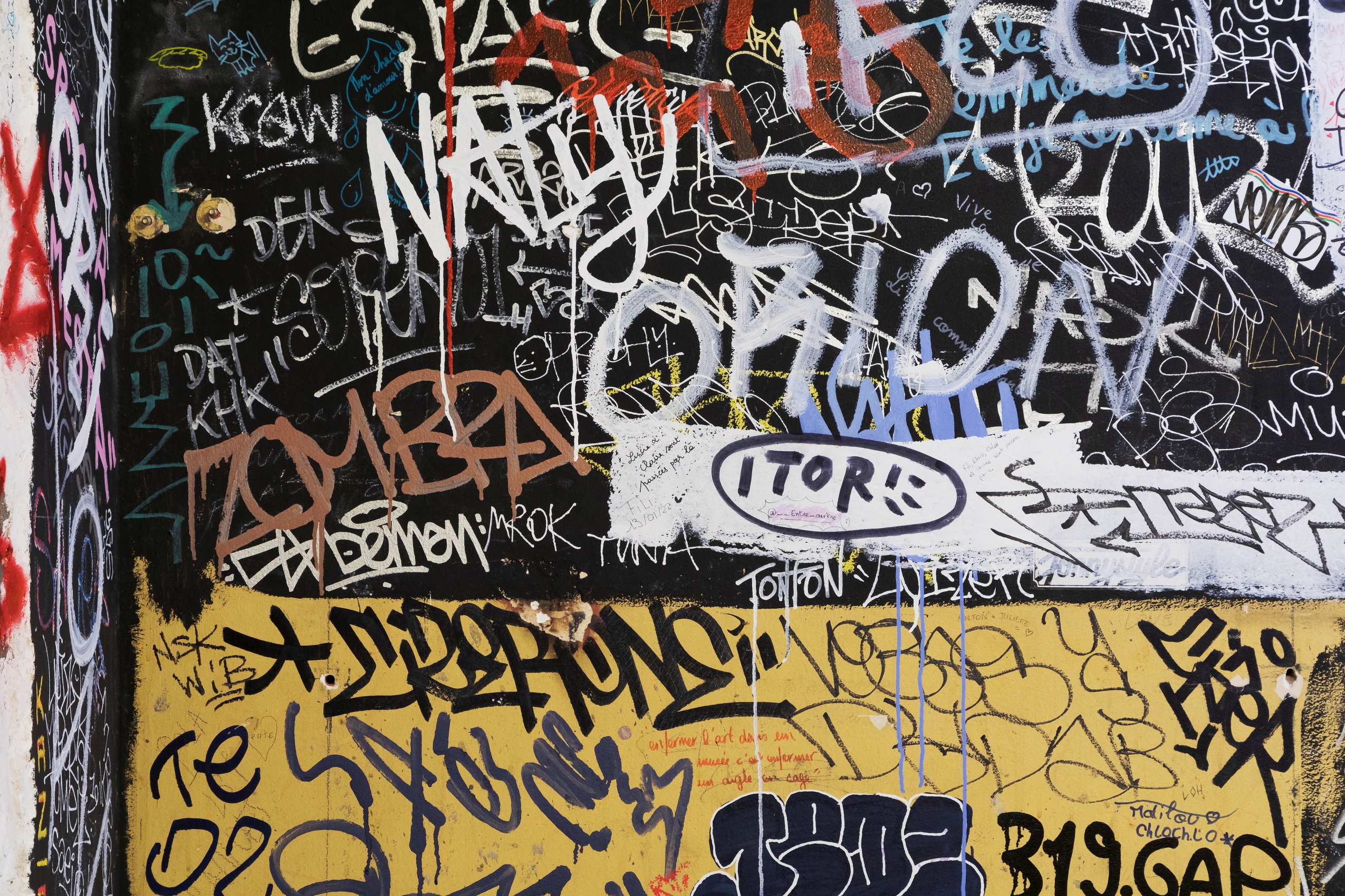 La portion de mur capturée sur l’image est séparée en deux sections colorées (noire et jaune). Elles sont équitablement recouvertes de graffitis plus ou moins amateurs : Zombra, Naty, Krow, Orion, Tonton, Demon, etc.