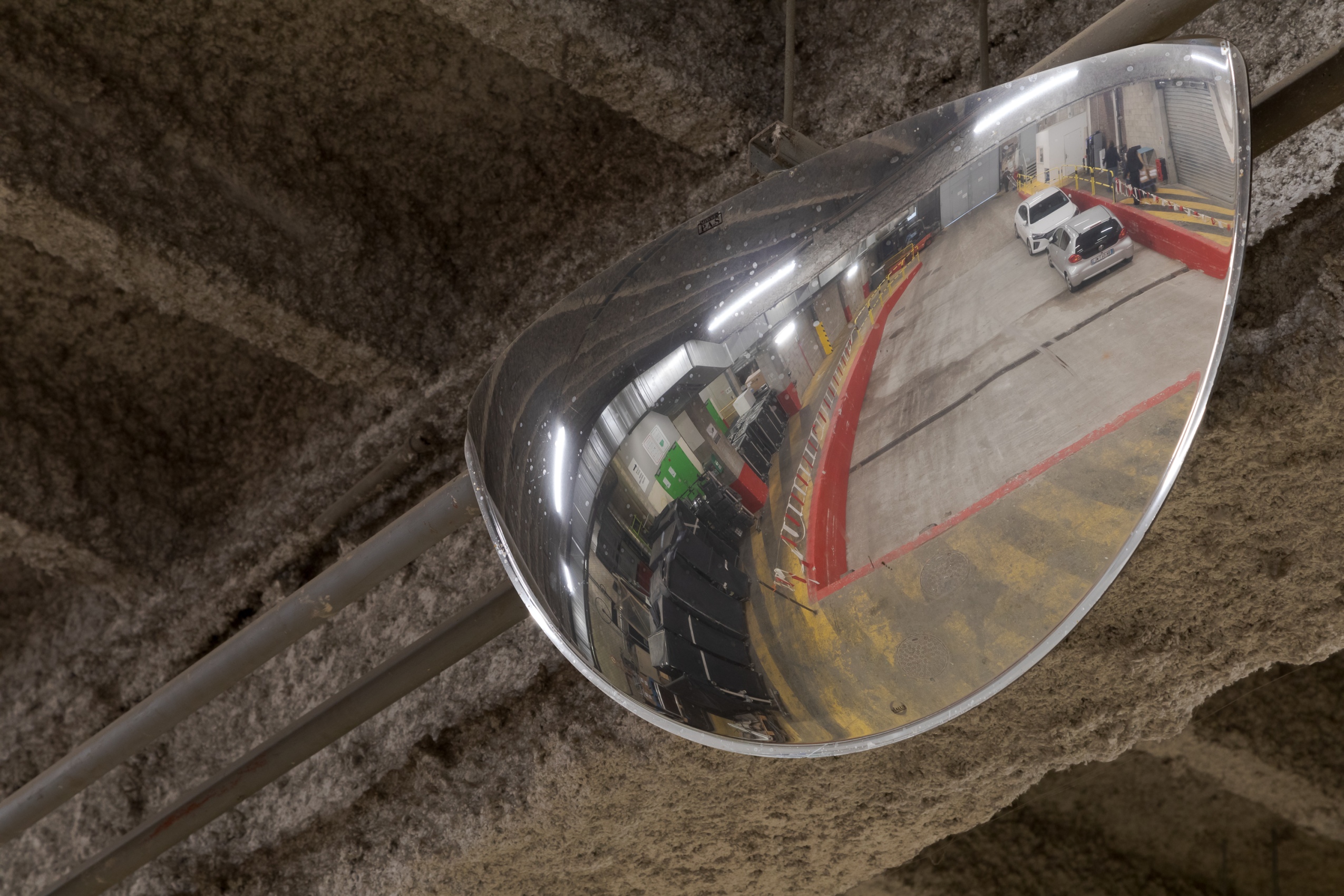 Un miroir courbe en métal est suspendu aux poutres floquées du quai de déchargement : on aperçoit dans le miroirs les lignes jaunes de la signalétique au sol ainsi que plusieurs voitures garées.