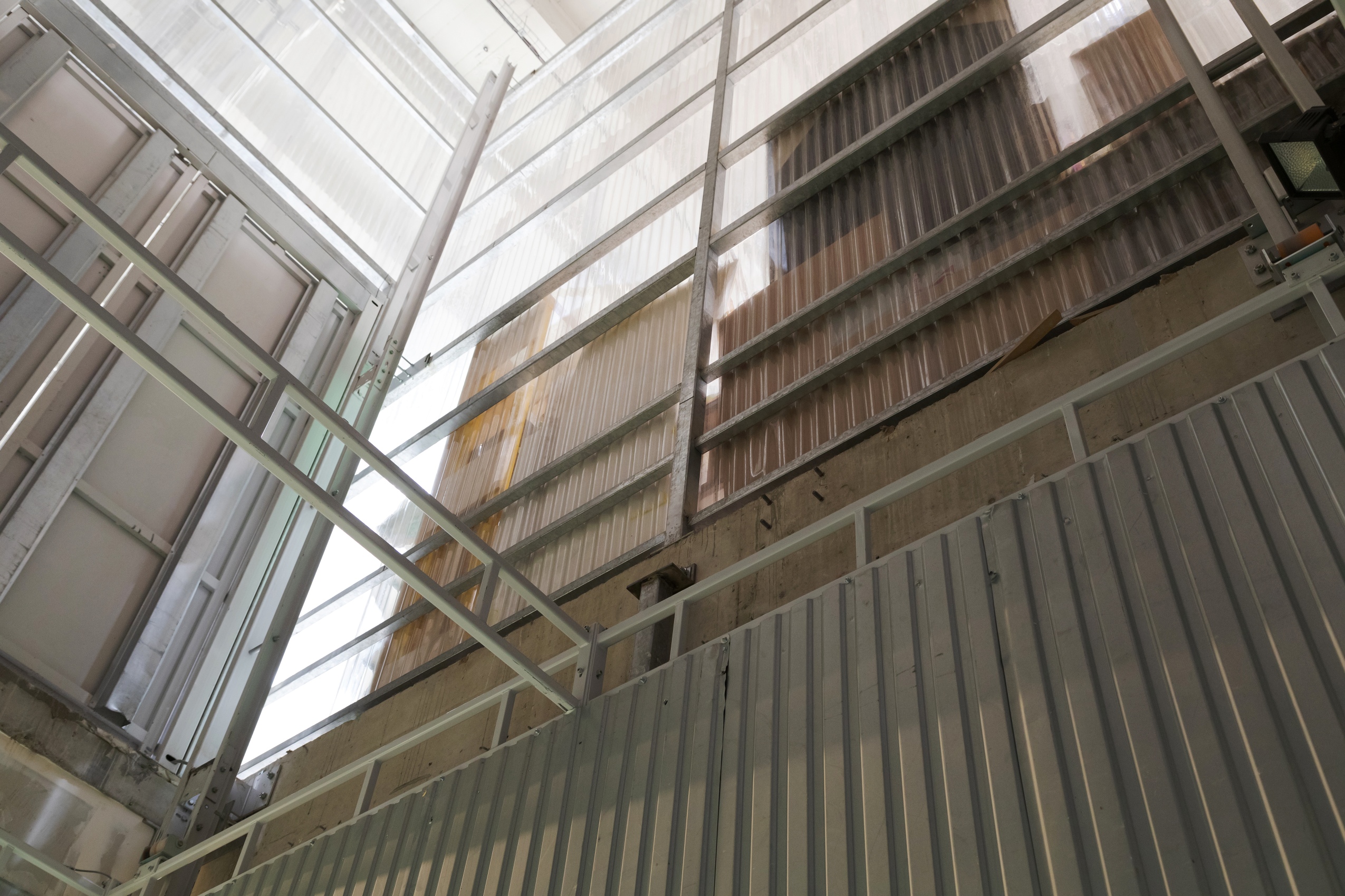 En contre plongée depuis la cage du monte charge, entouré de murs de tôles en métal puis en plastique transparent, on aperçoit l’étage supérieur, celui qui dessert les espaces d’exposition.
