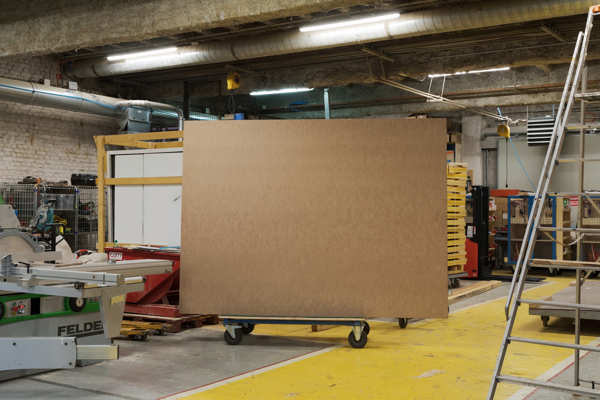 Dans l’un des ateliers du Palais de Tokyo (l’atelier bois), un grand panneau de médium brun est stationné au centre de l’espace, perché sur un chariot roulant. Autour, des machines en métal, des structures en bois, des palettes entreposées ou des échelles. 