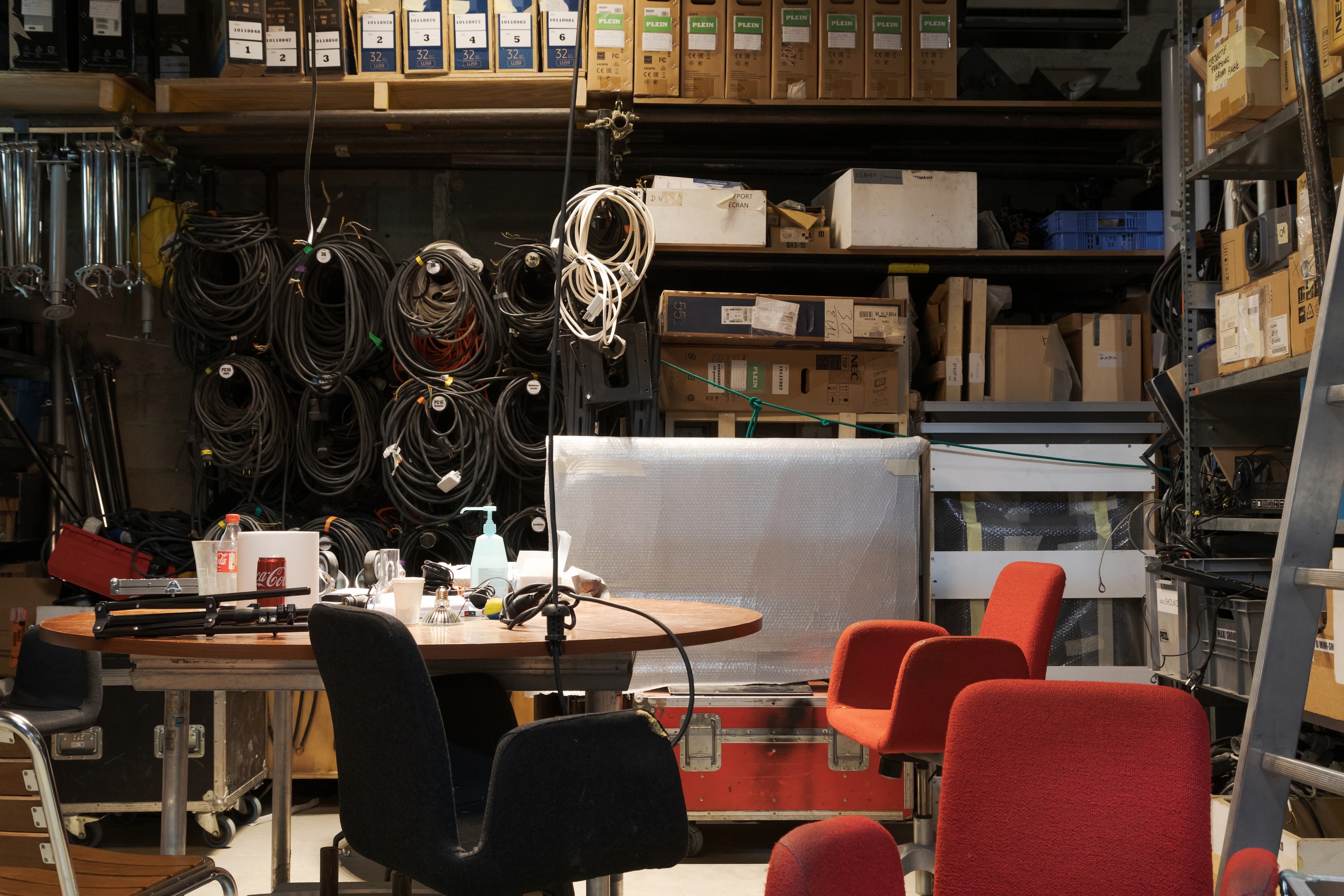 L’atelier de la régie audiovisuelle est organisé autour d’une table centrale, entouré de fauteuils rouges et noirs. Les murs sont couverts de câbles, de projecteurs, de rouleaux de plastique coloré, d’échelles ou de cartons.