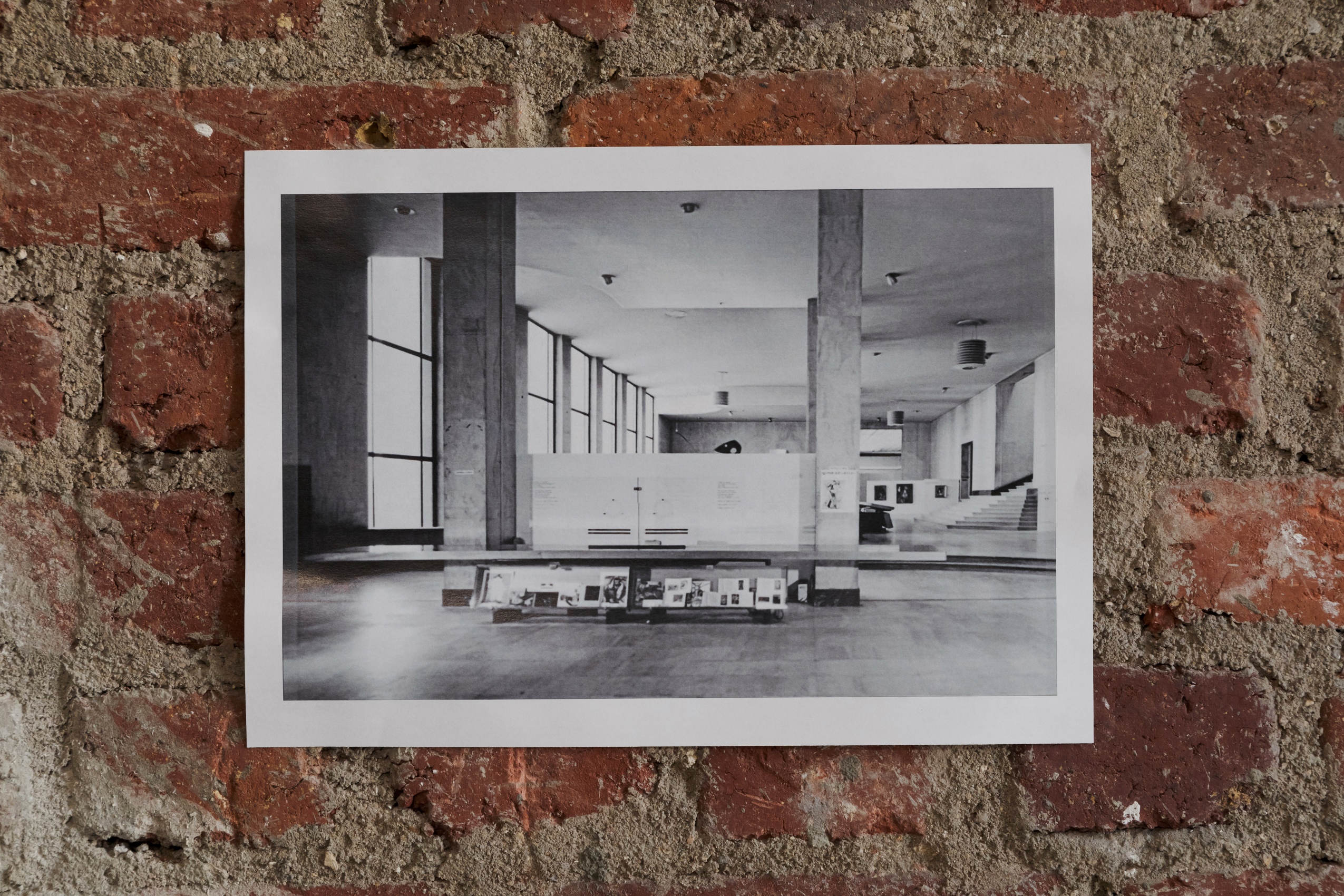 Sur un mur de briques rouges, un tirage d’une photographie datant de 1963 : la banque d’accueil en bois conçue par Charlotte Perriand, disposée entre deux colonnes. Des prospectus sont disposés en dessous de cette banque. 