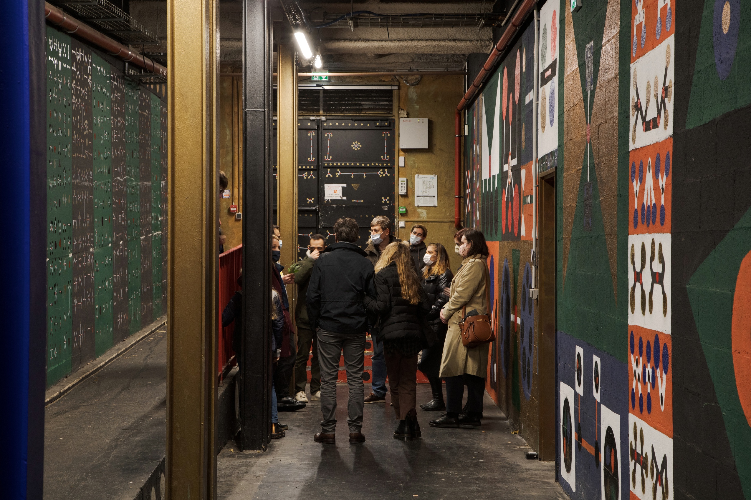 Un groupe de visiteurs entourent un médiateur culturel qui raconte l’histoire d’une fresque murale qui s’étend de part et d’autres : des symboles géométriques vert sapin, orangés, blanc et bleu klein. 