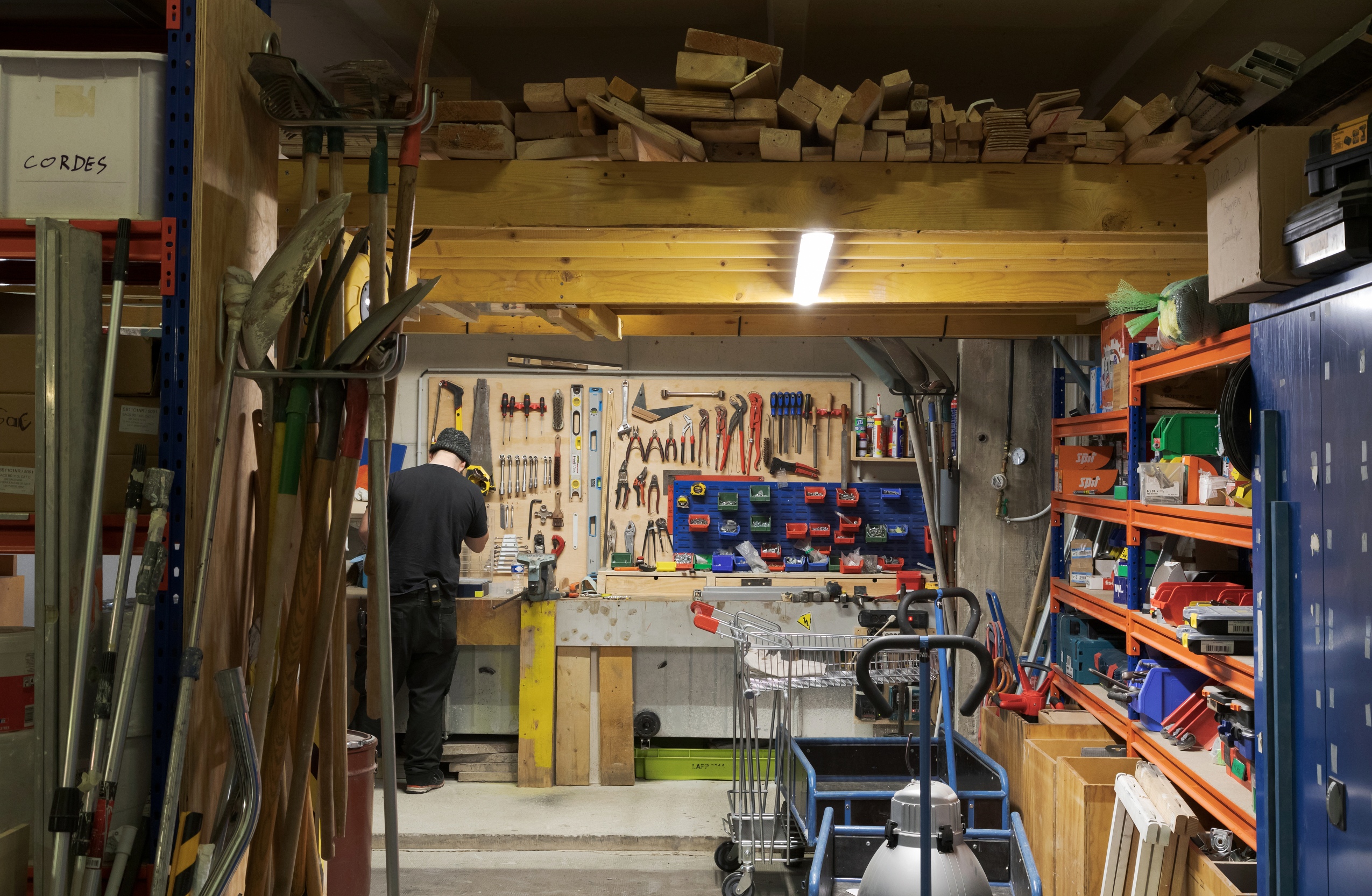 Dans l’atelier destiné aux réparations du bâtiment, on aperçoit Nicolas devant son établi : un mur ou chaque outil a sa place (pinces, marteaux, tournevis). Au plafond, des tasseaux de bois sont stockés tandis que sur les autres murs, on retrouve des pelles, des tréteaux, des tuyaux en plastique ou des chariots. 