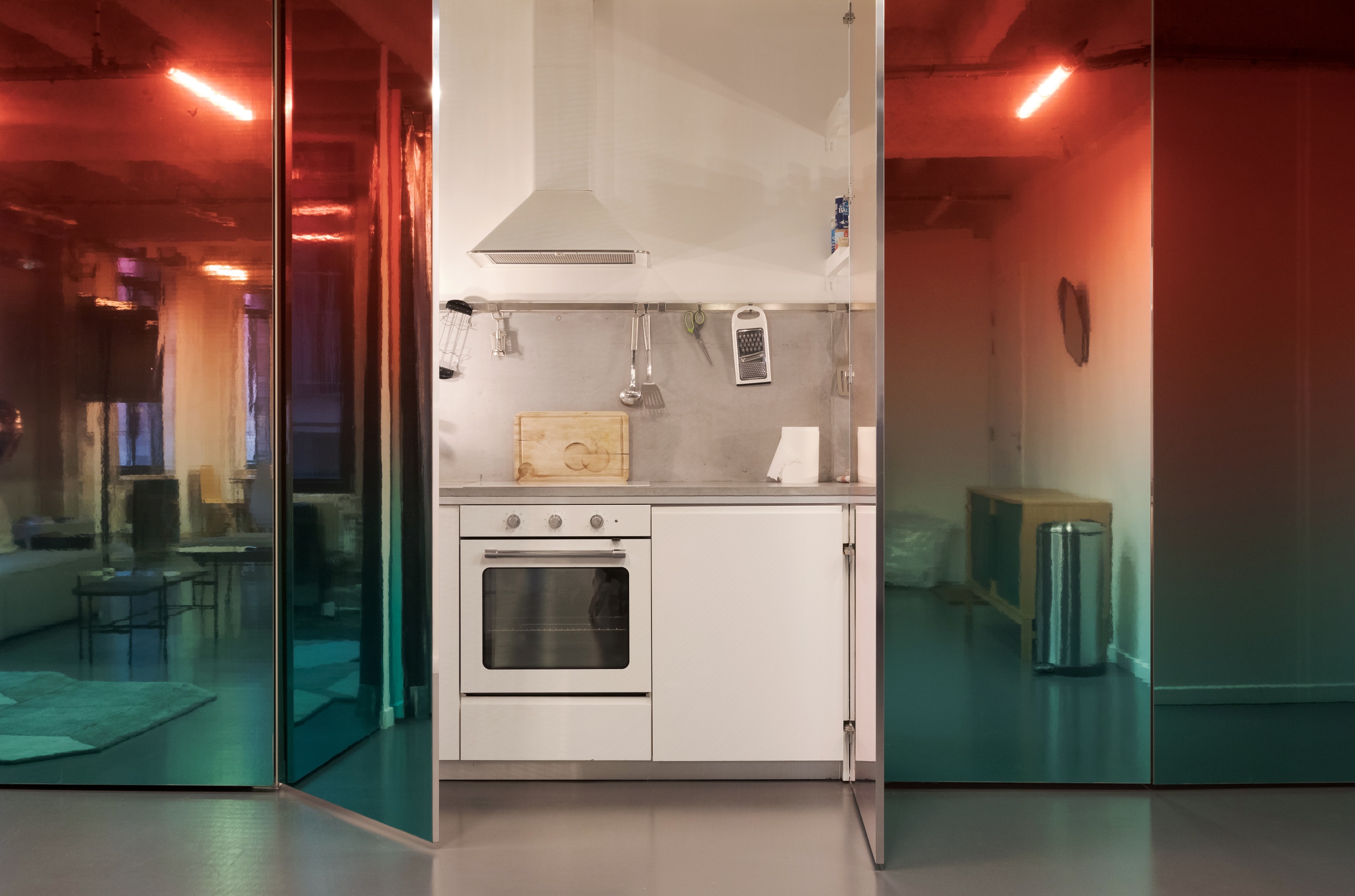 L’appartement des artistes est un espace brut dans lequel ont été conçus des modules fonctionnels (cuisine, chambre, salle de bain). Ces éléments sont masqués derrière un grand mur recouvert d’un miroir teinté d’un dégradé allant du rouge au bleu. Au centre, se dévoile une petite cuisine toute équipée.  