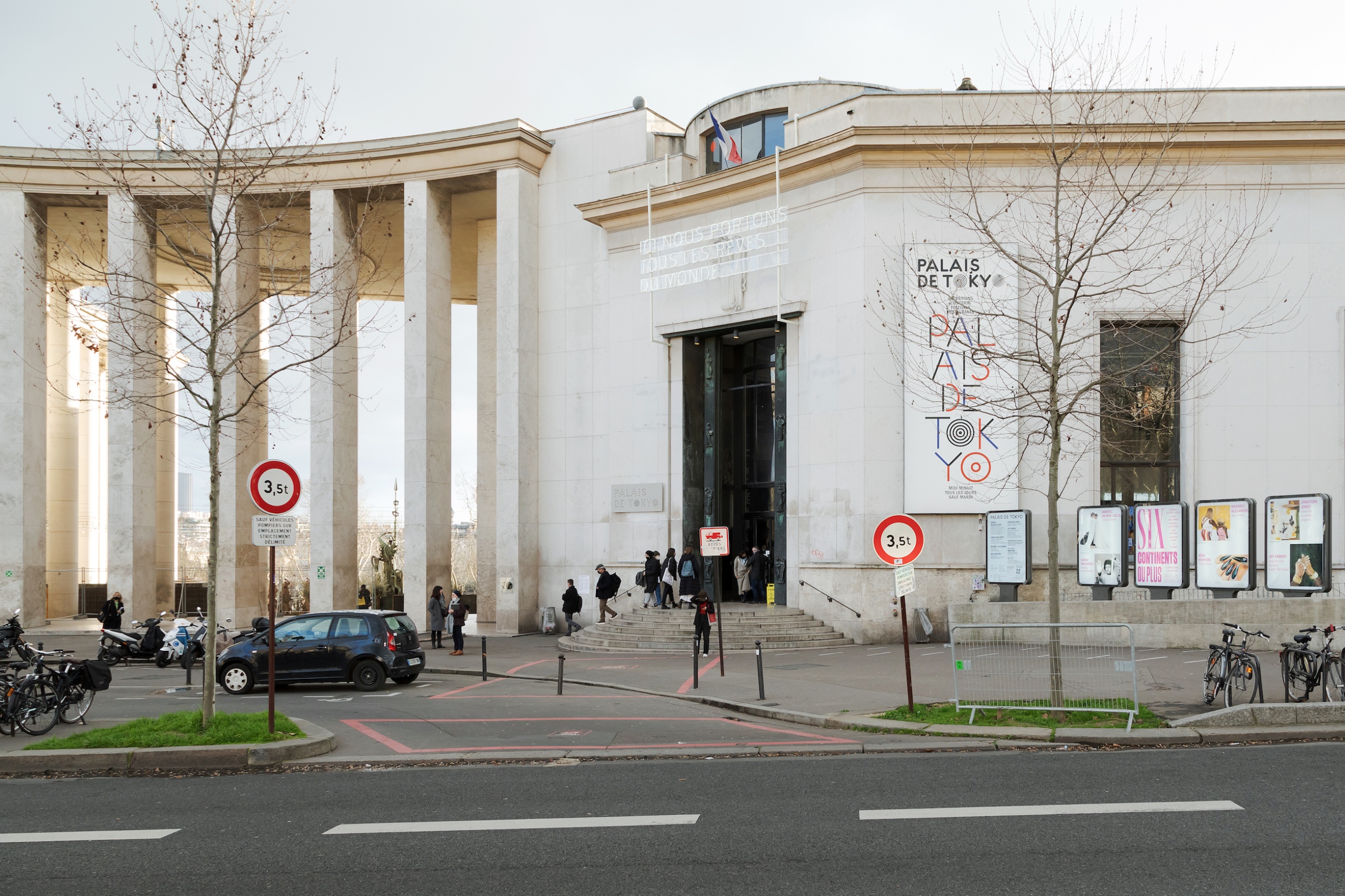 Le long de l’avenue du Président Wilson, le parvis marquant l’entrée du Palais de Tokyo voit se dresser une série incurvée de colonnes rectangulaires blanches. La porte d’entrée sombre voit défiler quotidiennement des centaines de visiteurs.