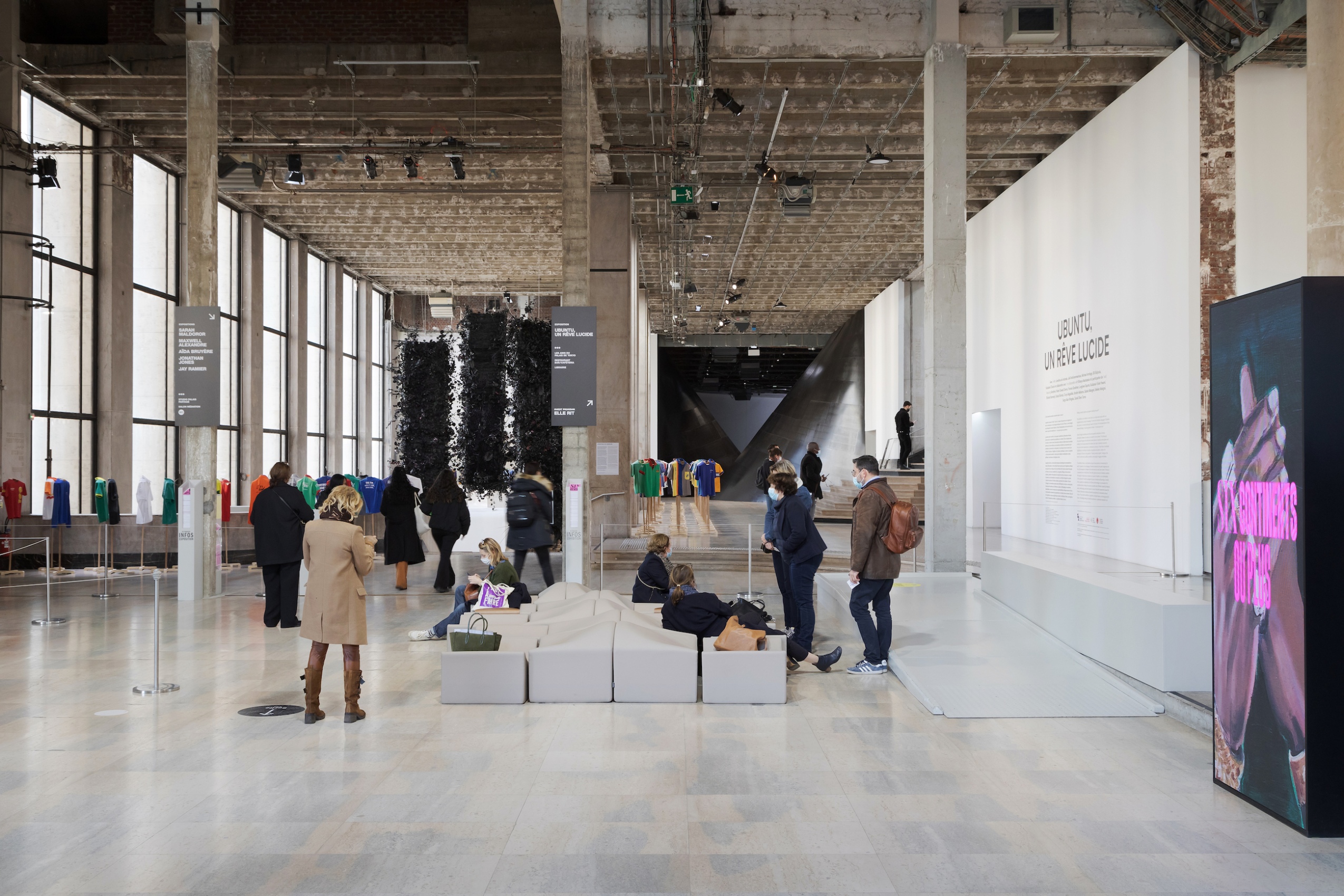 Les poutres en béton brut du plafond, l’enfilade des grandes fenêtres et les colonnes rectangulaires découpent le grand hall en de multiples images à explorer. Un espace d’exposition courbé dans l’ombre au fond, des œuvres suspendues à gauche, des escaliers à descendre et monter, monter et descendre sur la droite. 