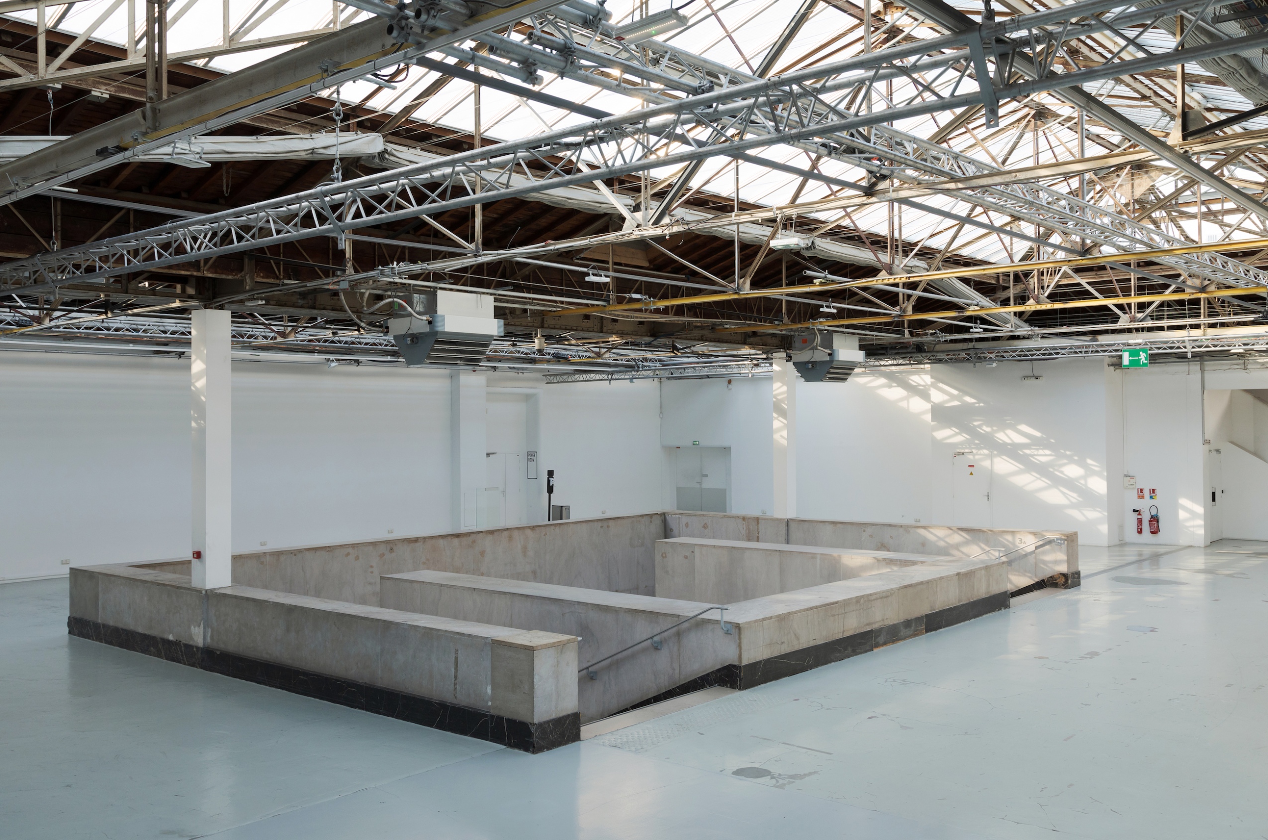 Sous les verrières, un grand hall vide (murs blancs et sol gris) est occupé en son centre par un grand escalier en pierre qui descend vers les espaces d’exposition. 